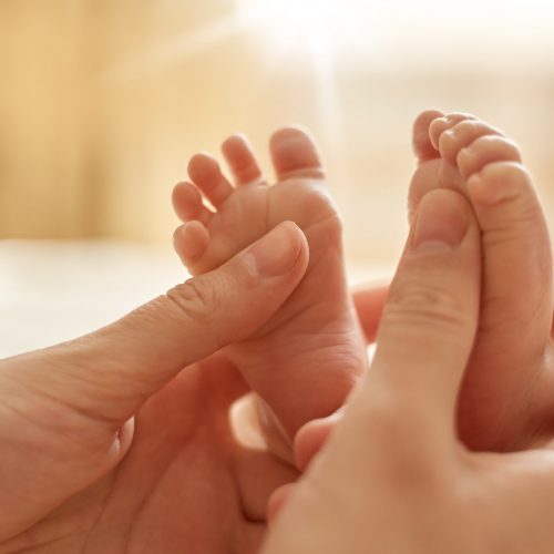 Mum making baby massage, mother massaging infant bare foot, preventive massage for newborn, mommy stroking the baby's feet with both hands on light background.